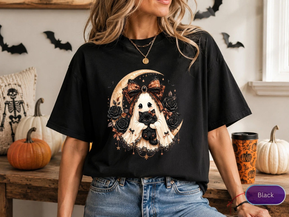 May include: A black t-shirt featuring a graphic print of a white ghost wearing a large brown patterned bow, holding a fluffy black kitten. The ghost is positioned in front of a crescent moon and surrounded by dark roses and lace-like accents.