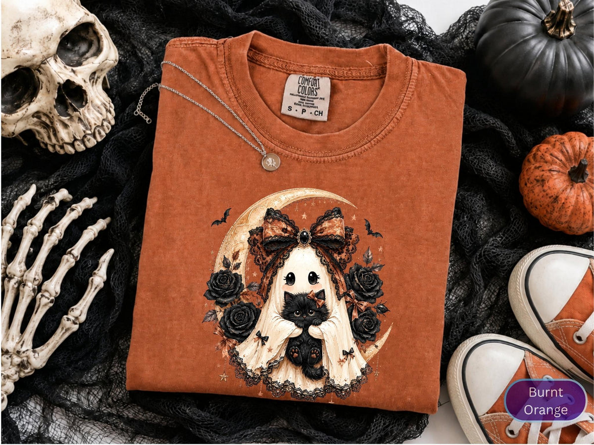May include: A burnt orange t-shirt featuring a graphic of a ghost wearing a large patterned bow and holding a black kitten. The design includes a crescent moon, black roses, and small bats. The shirt has a crew neckline and a Comfort Colors label.