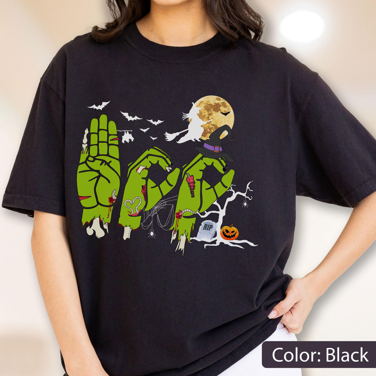 May include: A black t-shirt featuring a graphic of three green zombie hands forming the word BOO. The design includes a full moon, a witch on a broomstick, bats, a spider web, a tombstone with RIP text, and a jack-o'-lantern near a bare tree.