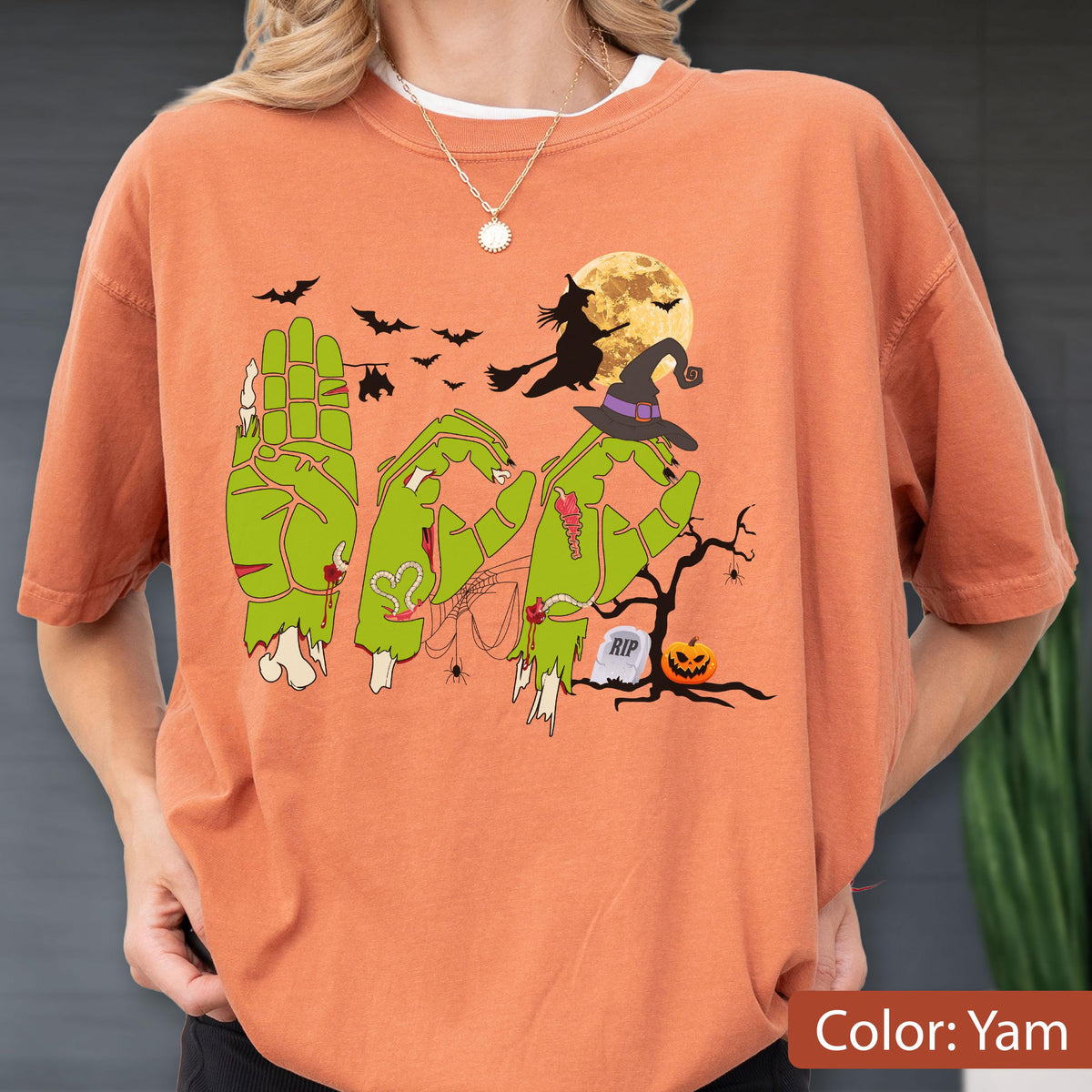 May include: A yam-colored t-shirt featuring a graphic of three green zombie hands forming the word BOO. The design includes a witch on a broomstick, a full moon, bats, a spider web, a tombstone with RIP text, and a jack-o'-lantern pumpkin.