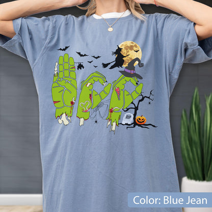 May include: A blue jean colored t-shirt featuring a graphic of three green zombie hands forming the letters B, O, and O. The design includes a witch on a broomstick, a full moon, bats, a spider web, a tombstone with RIP, and a jack-o'-lantern.