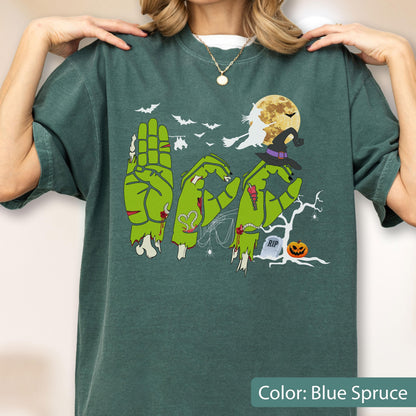 May include: A dark green t-shirt featuring a graphic of three green zombie hands forming the word BOO. The design includes a yellow full moon, a flying witch silhouette, bats, a witch hat, a bare tree, a tombstone with RIP text, and a jack-o'-lantern.