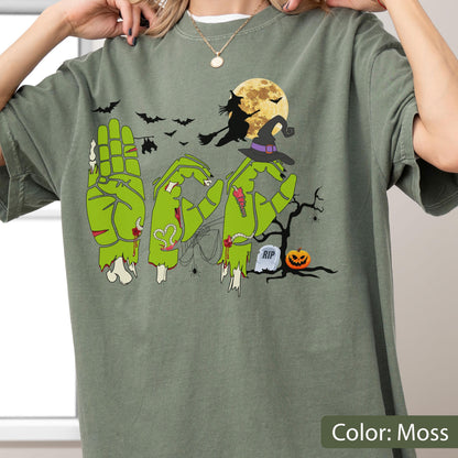 May include: A moss green t-shirt featuring a graphic of three green zombie hands forming the letters B-O-O in American Sign Language. The design includes a witch on a broomstick, a full moon, bats, a spider web, a tombstone with RIP, and a pumpkin.