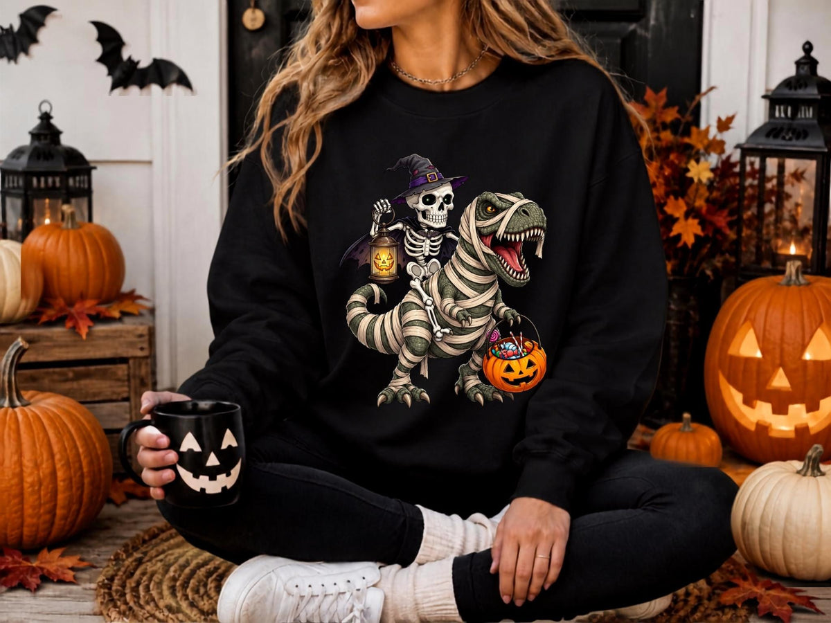May include: A black long-sleeve sweatshirt featuring a graphic of a skeleton wearing a purple witch hat and cape, riding a mummy-wrapped Tyrannosaurus Rex. The skeleton holds a glowing jack-o'-lantern lantern, and the dinosaur carries a candy bucket.