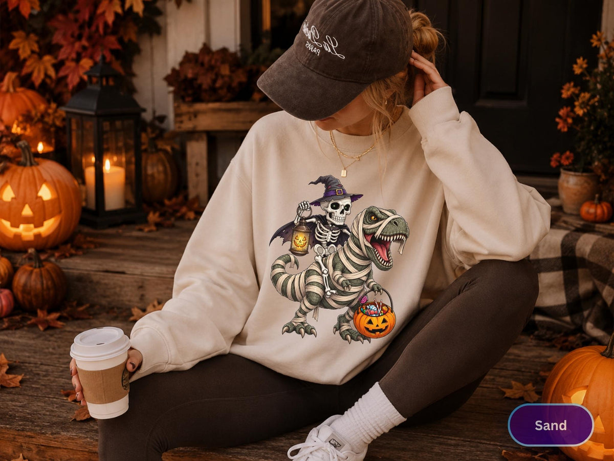 May include: A sand-colored crewneck sweatshirt featuring a graphic of a skeleton wearing a witch hat and cape, riding a mummy-wrapped Tyrannosaurus Rex. The skeleton holds a lantern, and the dinosaur carries a jack-o'-lantern bucket filled with candy.