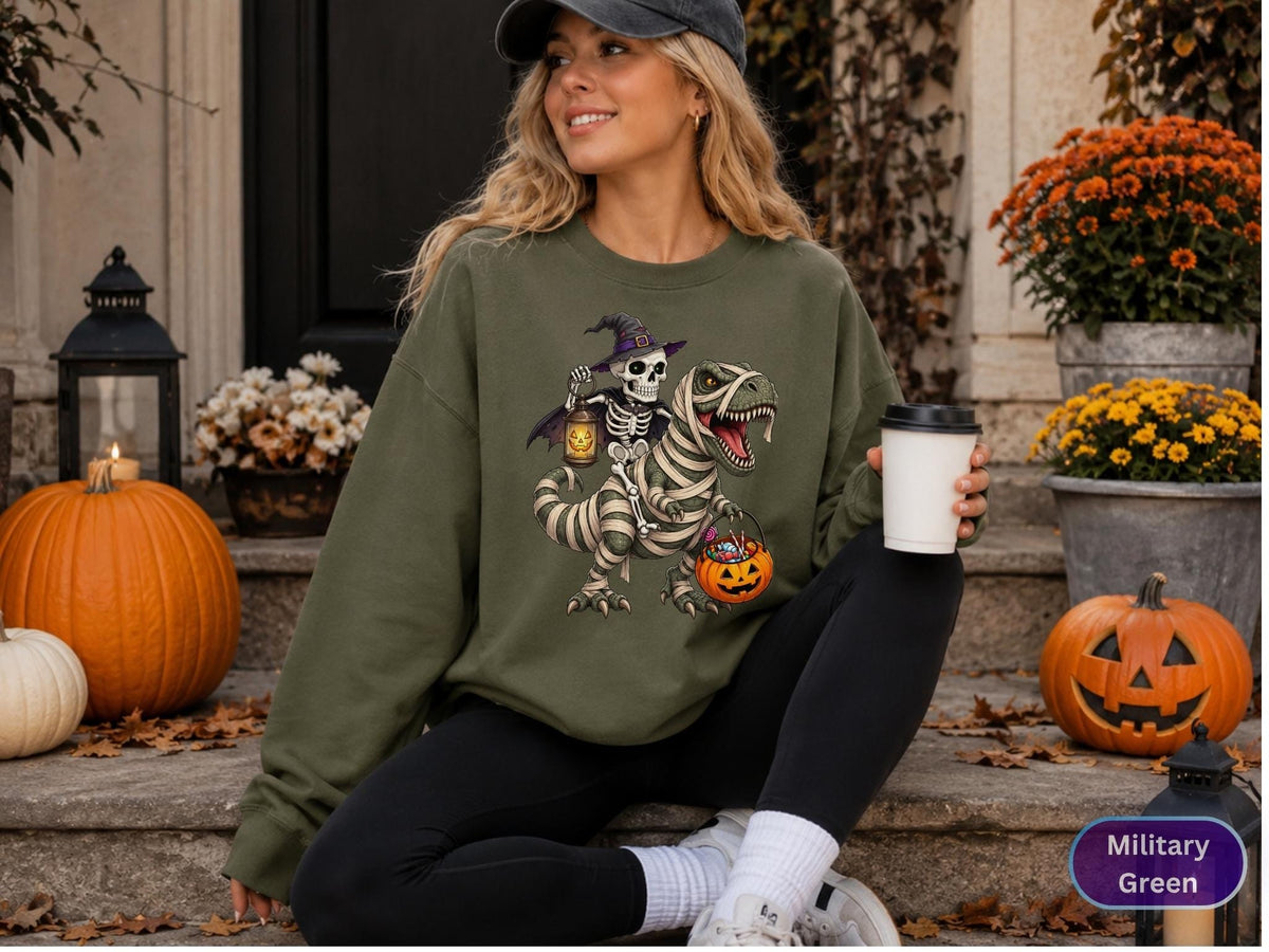May include: A military green crewneck sweatshirt featuring a graphic print of a skeleton wearing a witch hat and holding a lantern while riding a mummified Tyrannosaurus Rex that is carrying a jack-o'-lantern bucket filled with candy.