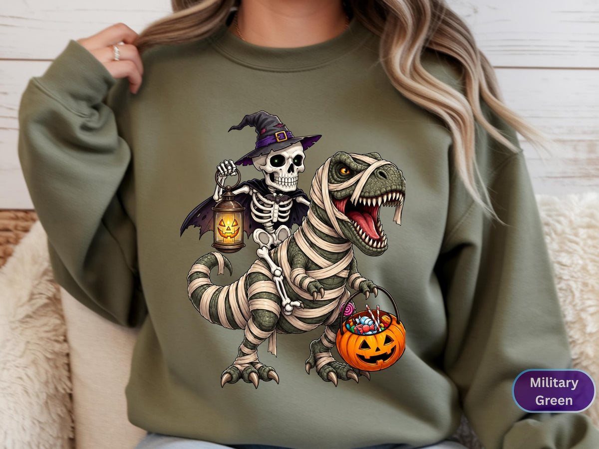 May include: A military green crewneck sweatshirt featuring a graphic of a skeleton wearing a witch hat and cape, holding a lit jack-o'-lantern lantern while riding a T-Rex wrapped in mummy bandages and carrying a pumpkin bucket filled with candy.