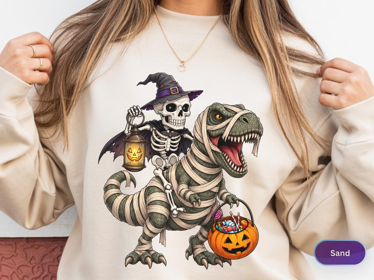 May include: A sand-colored sweatshirt featuring a graphic print of a skeleton wearing a purple witch hat and cape, holding a glowing jack-o'-lantern lantern while riding a green T-Rex wrapped in white mummy bandages and carrying a pumpkin candy bucket.