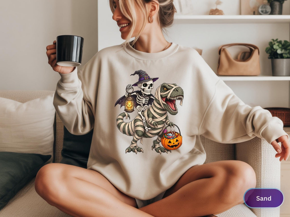 May include: A sand-colored crewneck sweatshirt featuring a graphic print of a skeleton wearing a purple witch hat and cape, riding a mummy-wrapped Tyrannosaurus Rex. The skeleton holds a glowing lantern, and the dinosaur carries a jack-o'-lantern bucket.