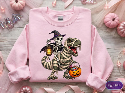 May include: A light pink crewneck sweatshirt featuring a graphic print of a skeleton wearing a witch hat and holding a lantern while riding a mummy-wrapped Tyrannosaurus Rex that is carrying a jack-o'-lantern bucket filled with colorful candy.