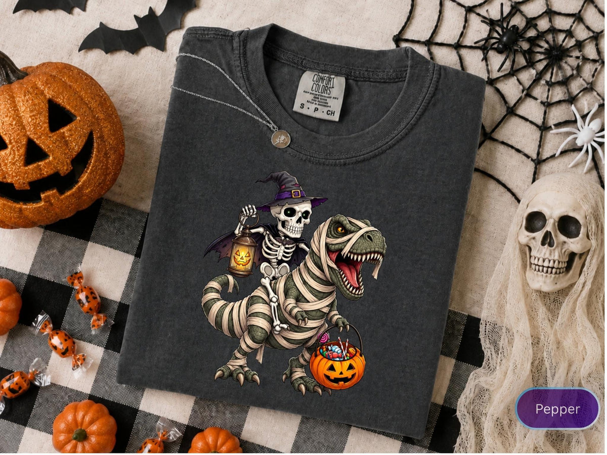 May include: A charcoal gray crew neck t-shirt featuring a graphic of a skeleton wearing a purple witch hat and holding a glowing lantern while riding a mummy-wrapped Tyrannosaurus Rex that is carrying a jack-o'-lantern bucket filled with candy.