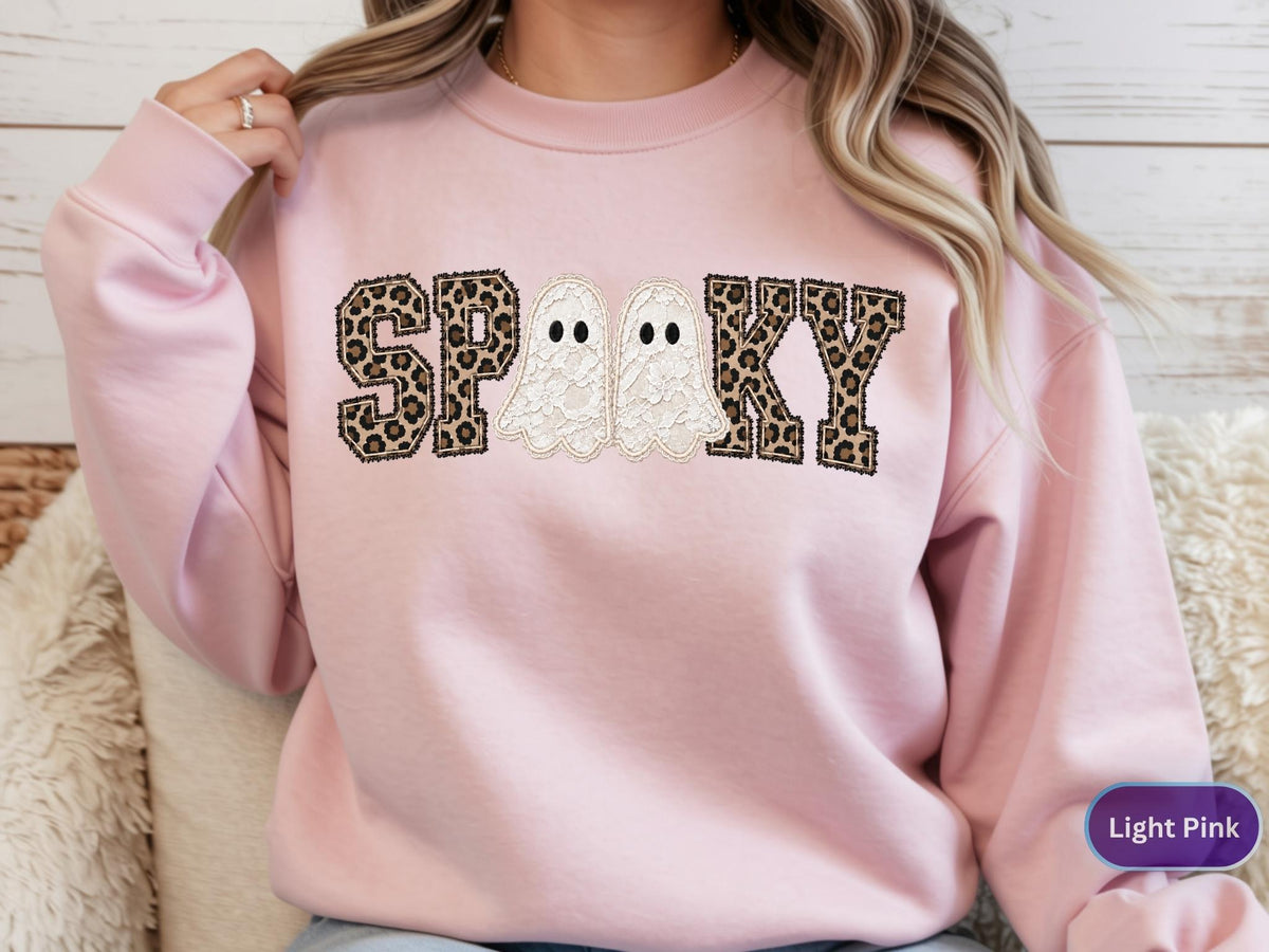 May include: A light pink crewneck sweatshirt featuring the word SPOOKY in a block font. The letters S, P, O, K, and Y have a brown and black leopard print pattern. Two white ghost figures with black eyes replace the letter O in the center.