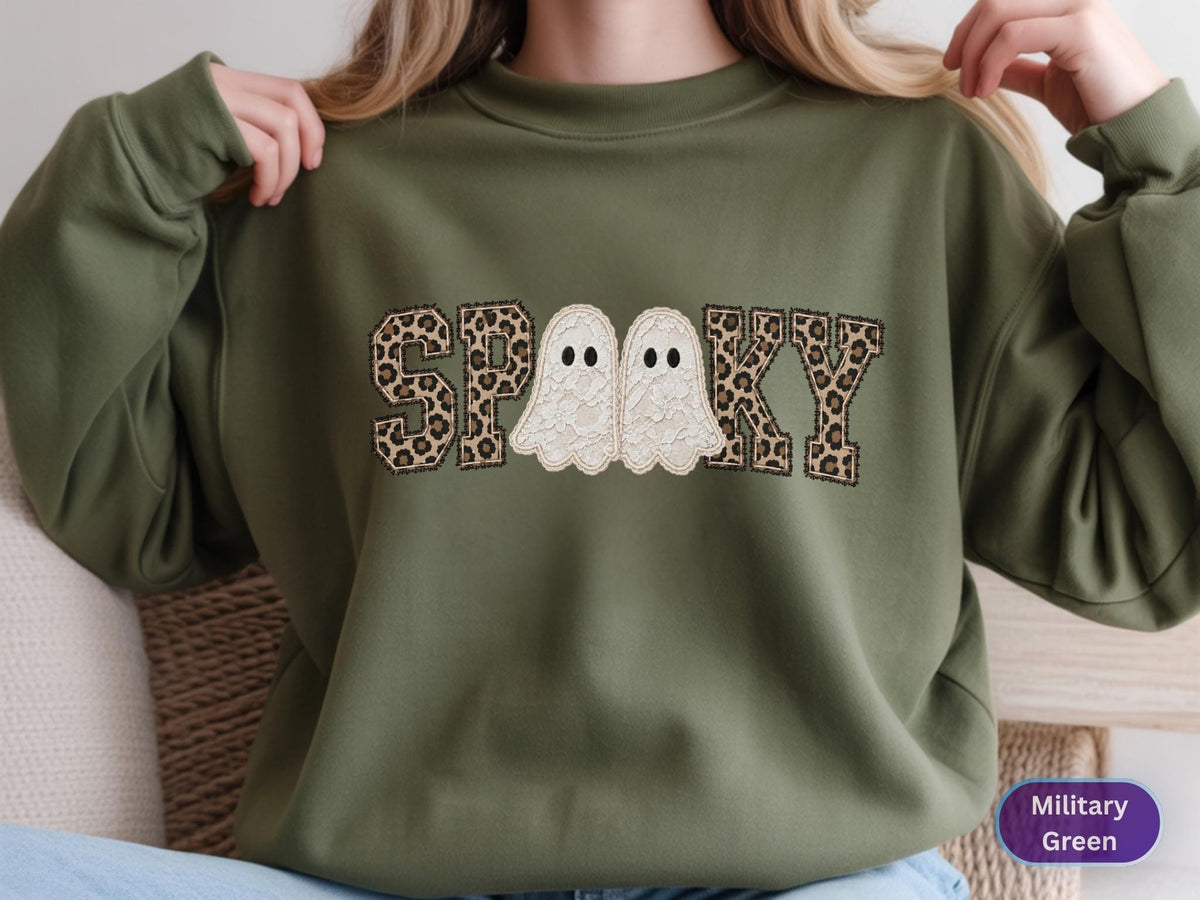 May include: A military green crewneck sweatshirt featuring the word SPOOKY embroidered across the chest. The letters S, P, K, and Y have a brown and black leopard print pattern, while the two O's are replaced by white ghost figures.