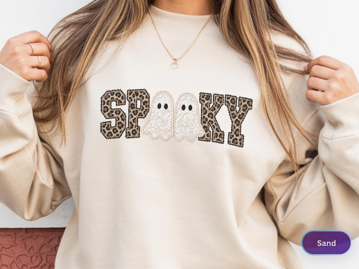 May include: A sand-colored crewneck sweatshirt featuring the word SPOOKY printed across the chest. The letters S, P, K, and Y have a brown and black leopard print pattern, while the two O's are replaced by white ghost illustrations.