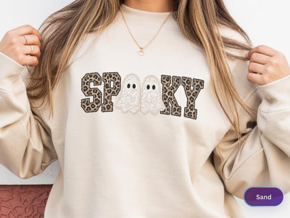 May include: A sand-colored crewneck sweatshirt featuring the word SPOOKY printed across the chest. The letters S, P, K, and Y have a brown and black leopard print pattern, while the two O's are replaced by white ghost illustrations.