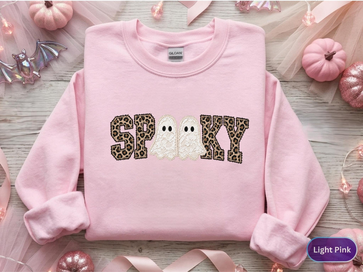 May include: A light pink crewneck sweatshirt featuring the word SPOOKY embroidered across the chest. The letters S, P, O, K, and Y are filled with a brown and black leopard print pattern, while two white ghost figures replace the letter O.