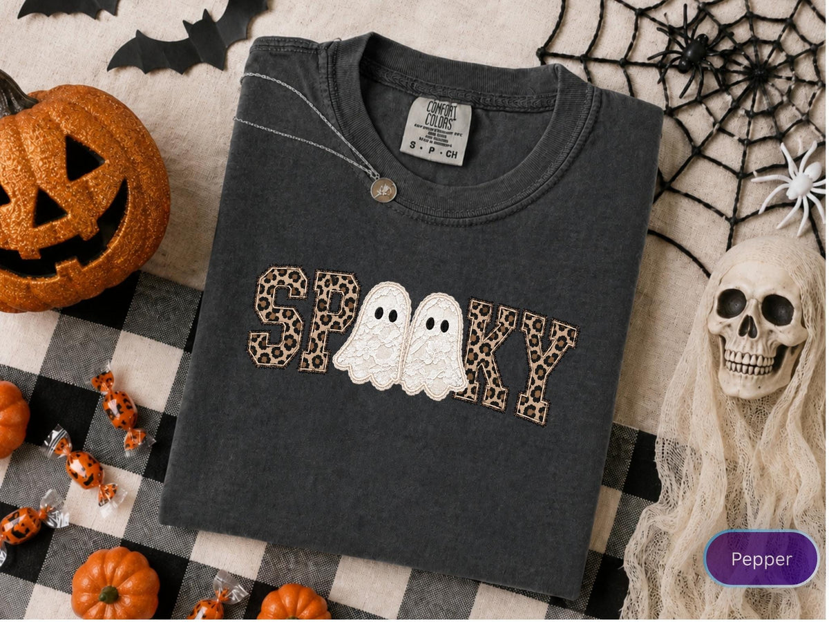 May include: A charcoal gray short-sleeve t-shirt featuring the word SPOOKY embroidered across the chest. The letters S, P, O, K, and Y are filled with a brown and black leopard print pattern, while two white lace ghosts replace the double O.