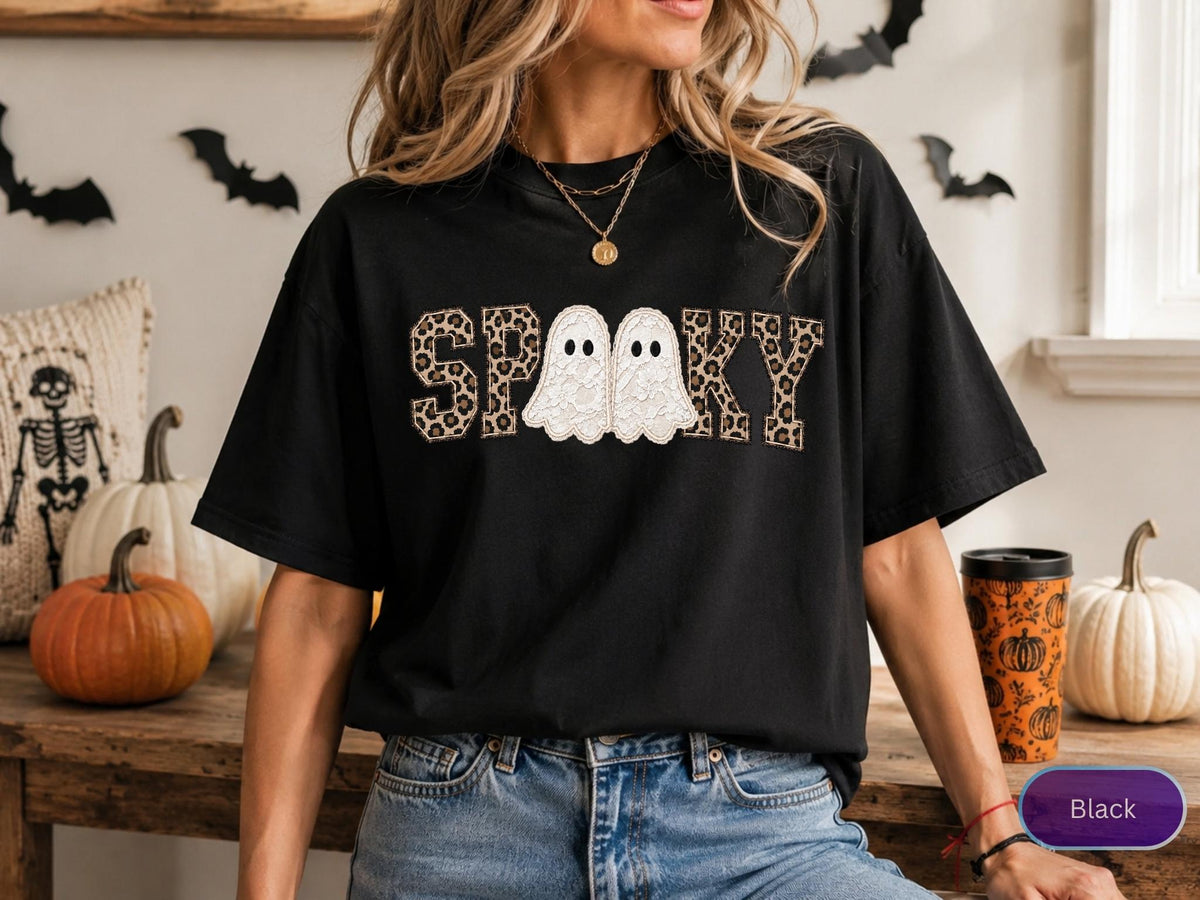 May include: A black short-sleeved t-shirt featuring the word SPOOKY printed across the chest. The letters S, P, O, K, and Y have a brown and black leopard print pattern, while the two O's are replaced by white ghost illustrations.