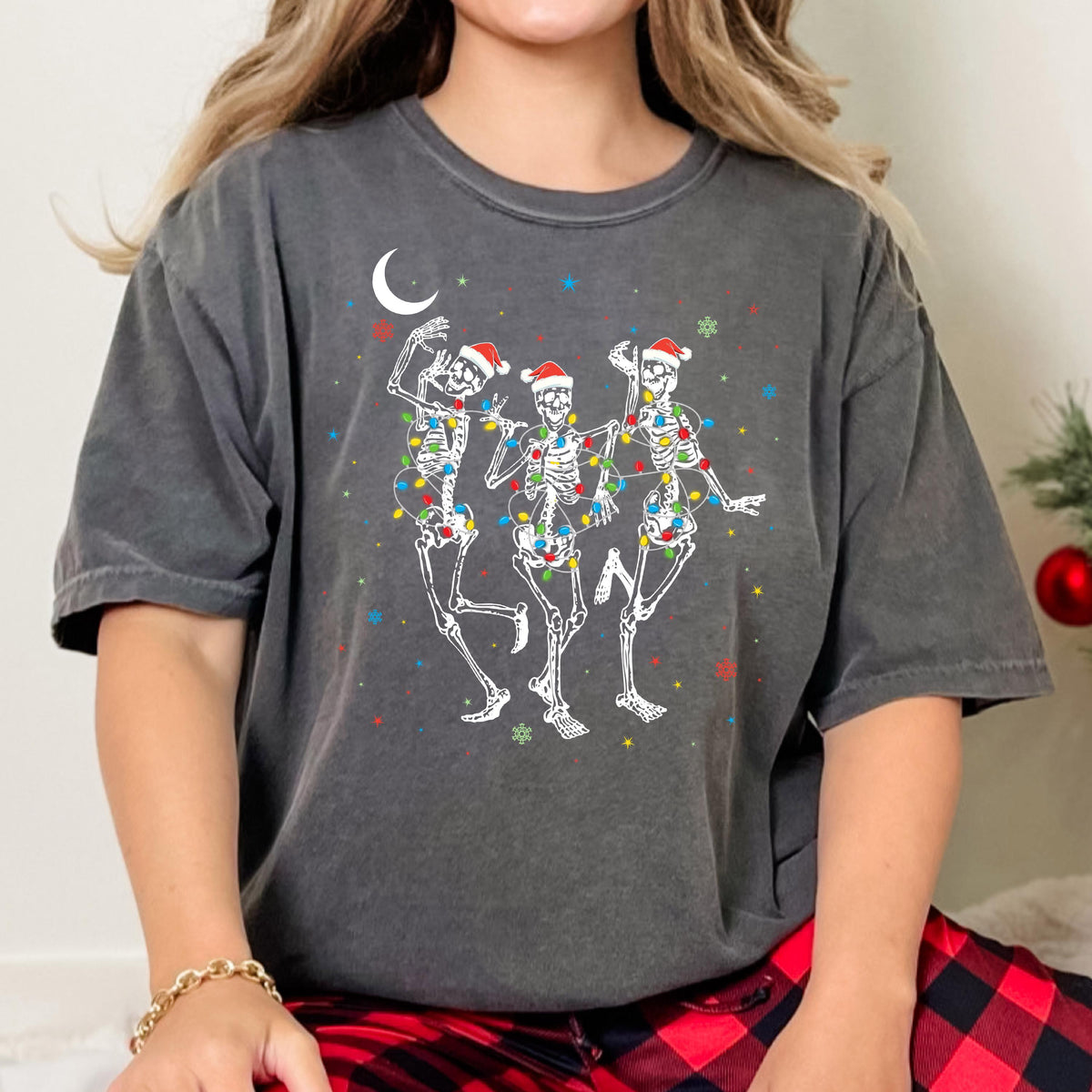 May include: A charcoal gray short-sleeve t-shirt featuring a graphic of three white skeletons wearing red Santa hats. The skeletons are tangled in colorful Christmas lights, with a white crescent moon and small colorful stars in the background.