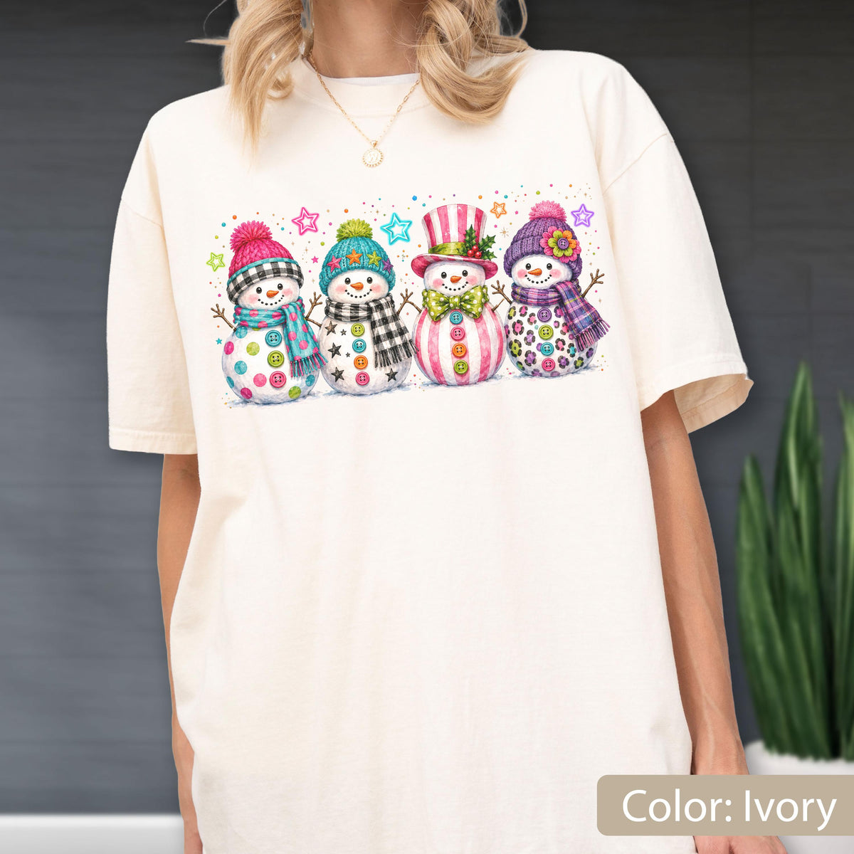 May include: An ivory t-shirt featuring a graphic print of four snowmen standing in a row. Each snowman wears a colorful winter hat and scarf with distinct patterns including plaid, polka dots, and leopard print, accented by small stars.