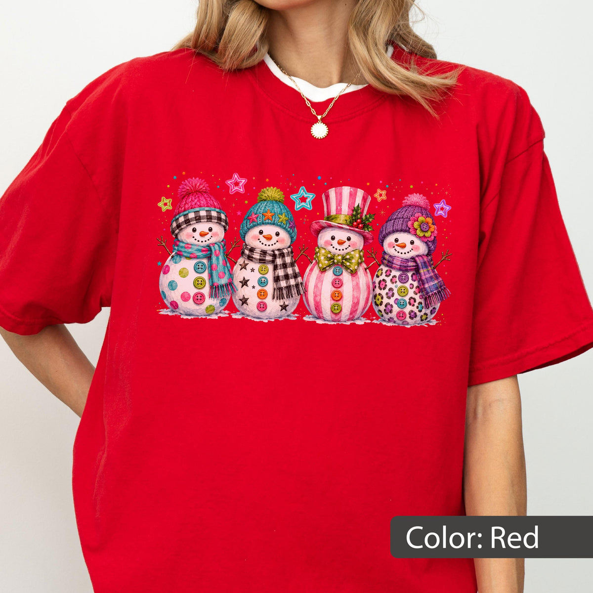 May include: A bright red short-sleeved t-shirt featuring a graphic print of four snowmen standing in a row. Each snowman wears a colorful hat and scarf, with various patterns like plaid, stars, and polka dots, and buttons on their bodies.