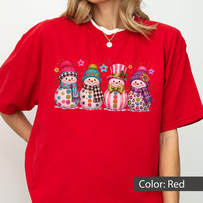 May include: A bright red short-sleeved t-shirt featuring a graphic print of four snowmen standing in a row. Each snowman wears a colorful hat and scarf, with various patterns like plaid, stars, and polka dots, and buttons on their bodies.