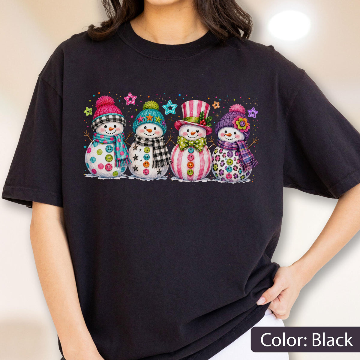 May include: A black t-shirt featuring a graphic print of four snowmen standing in a row. Each snowman wears a colorful hat and scarf with unique patterns, including plaid, stripes, and polka dots, accented by small stars scattered above them.