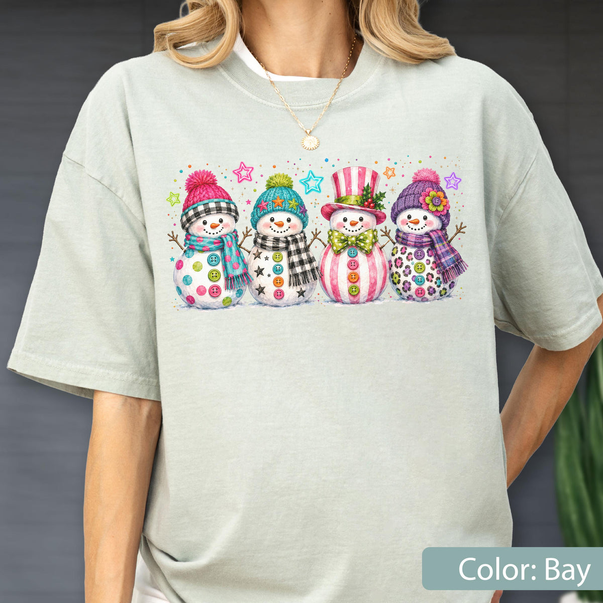 May include: A light sage green t-shirt featuring a graphic print of four snowmen standing in a row. Each snowman wears a colorful hat and scarf with unique patterns, including polka dots, plaid, and stripes, accented by small colorful stars.