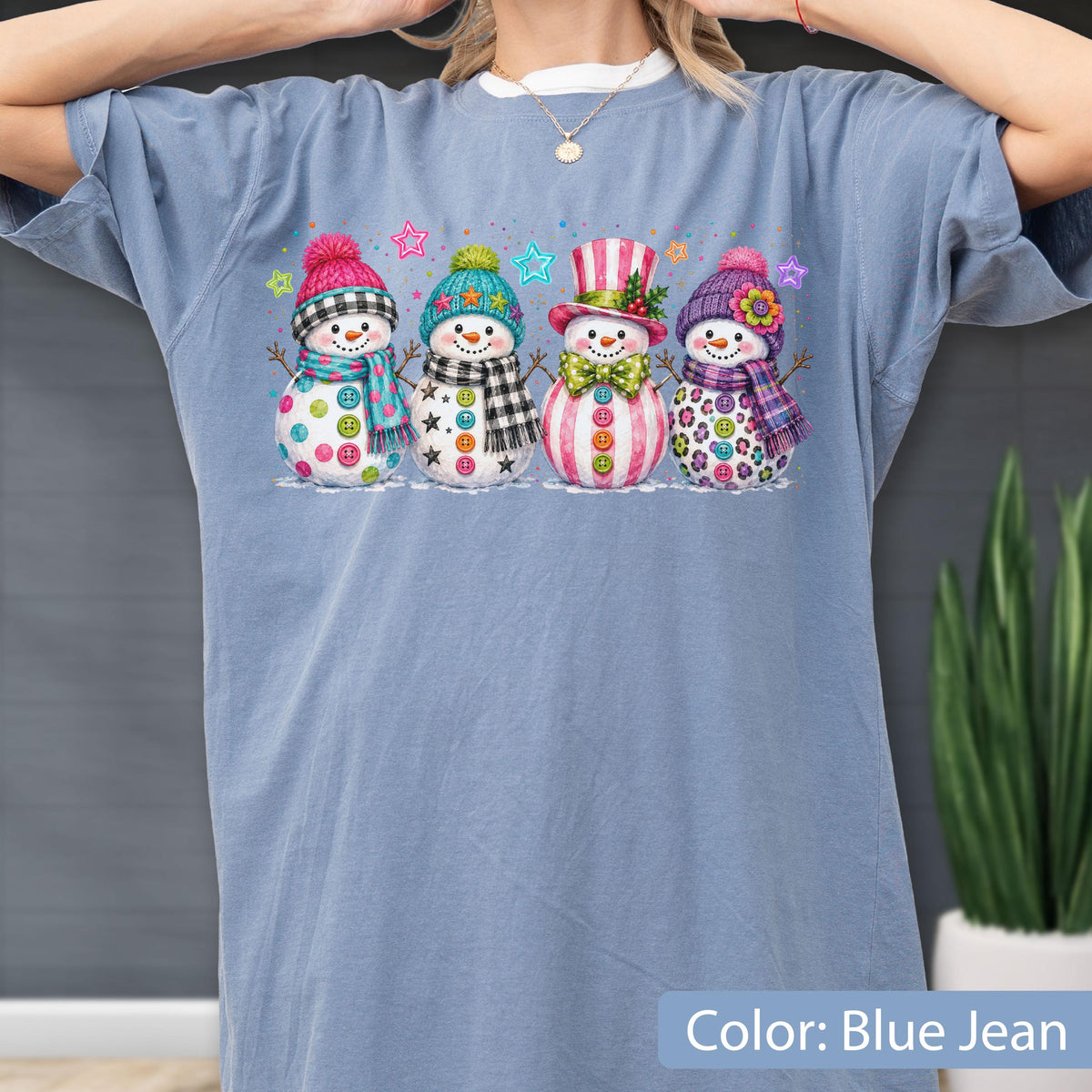 May include: A blue jean colored t-shirt featuring a graphic print of four snowmen standing in a row. Each snowman wears a different colorful hat and scarf, decorated with various patterns including buttons, stars, stripes, and floral accents.
