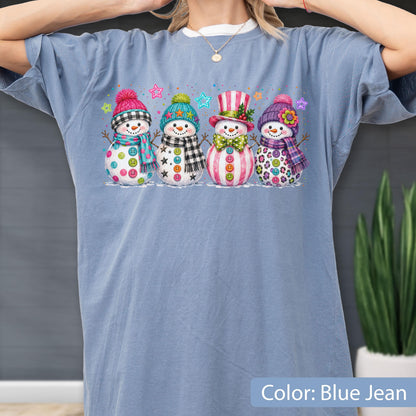 May include: A blue jean colored t-shirt featuring a graphic print of four snowmen standing in a row. Each snowman wears a different colorful hat and scarf, decorated with various patterns including buttons, stars, stripes, and floral accents.