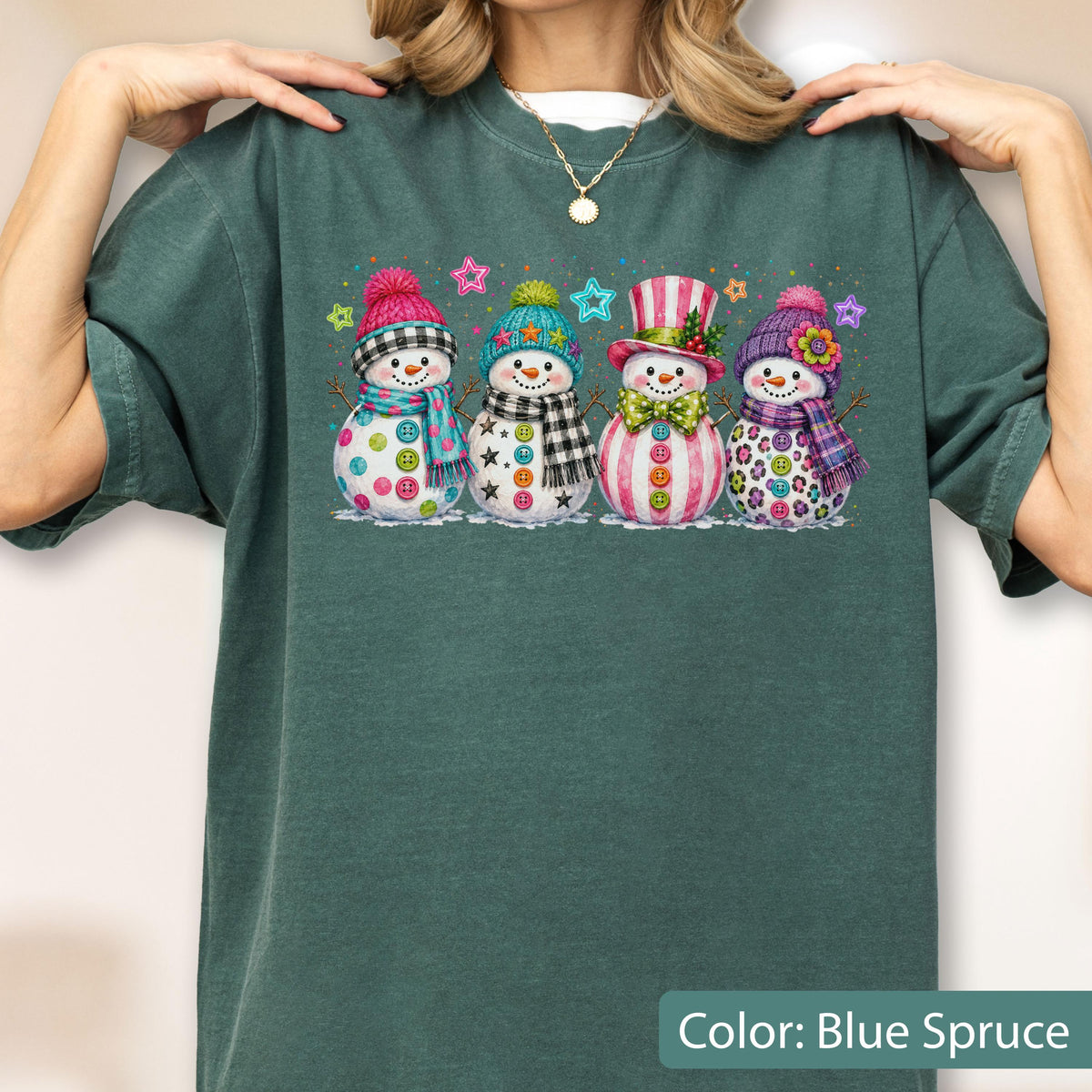 May include: A dark blue-green crewneck t-shirt featuring a graphic print of four snowmen wearing colorful hats, scarves, and patterned accessories. The snowmen are decorated with buttons and surrounded by small, multi-colored star shapes.