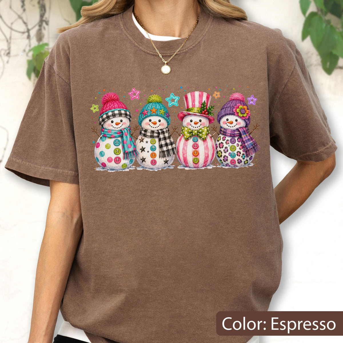 May include: An espresso-colored short-sleeve t-shirt featuring a graphic print of four snowmen standing in a row. Each snowman wears a colorful winter hat and scarf, with various button designs on their bodies and small star accents above them.