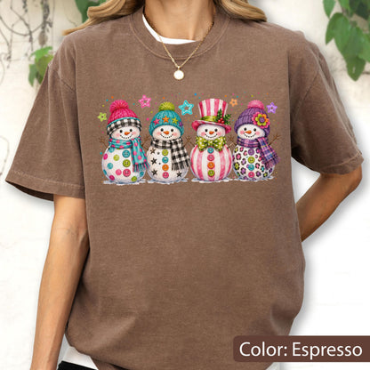 May include: An espresso-colored short-sleeve t-shirt featuring a graphic print of four snowmen standing in a row. Each snowman wears a colorful winter hat and scarf, with various button designs on their bodies and small star accents above them.