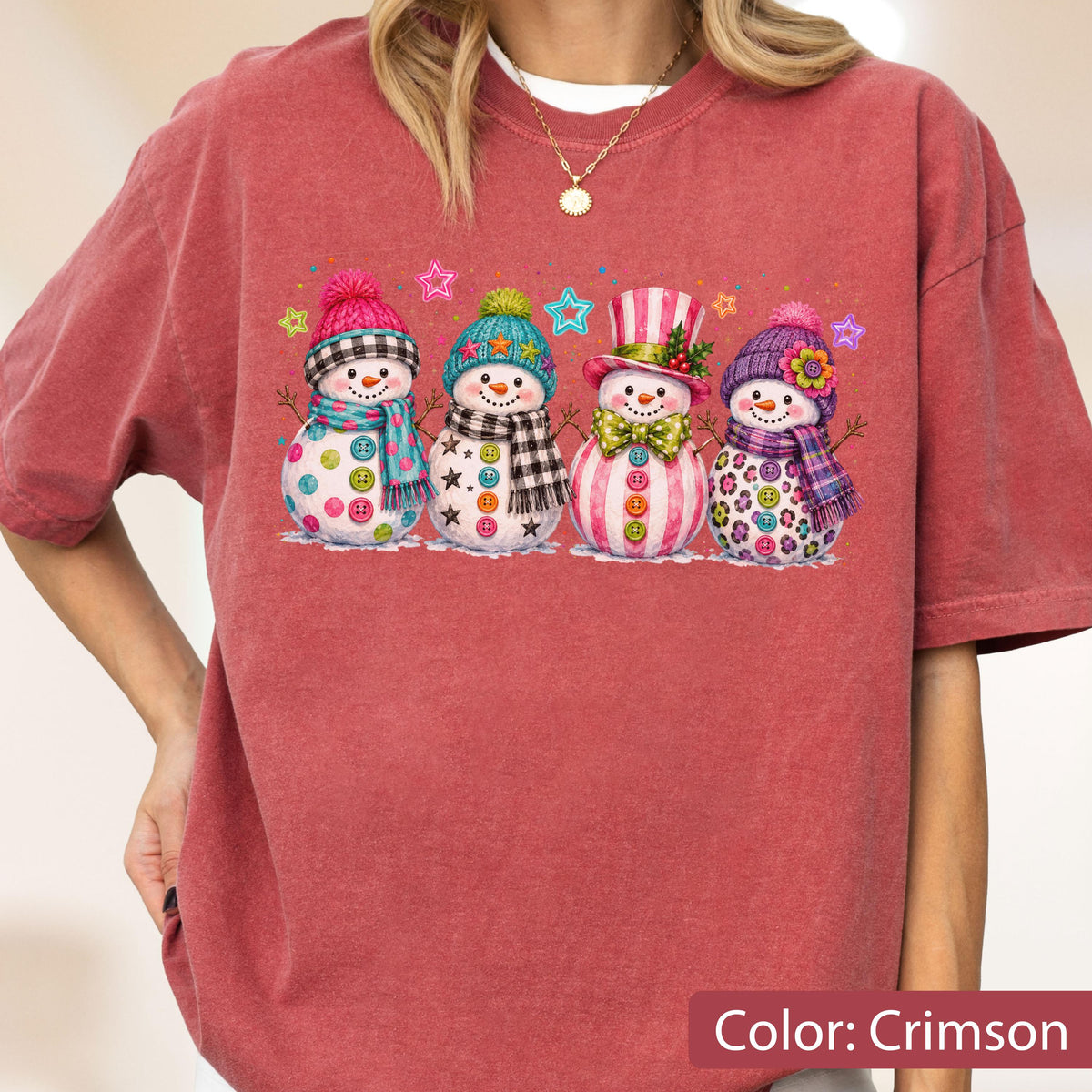 May include: A crimson short-sleeve t-shirt featuring a graphic print of four snowmen wearing colorful hats, scarves, and button accents. The snowmen are decorated with various patterns and surrounded by small, multi-colored star shapes.