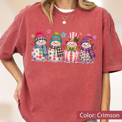 May include: A crimson short-sleeve t-shirt featuring a graphic print of four snowmen wearing colorful hats, scarves, and button accents. The snowmen are decorated with various patterns and surrounded by small, multi-colored star shapes.
