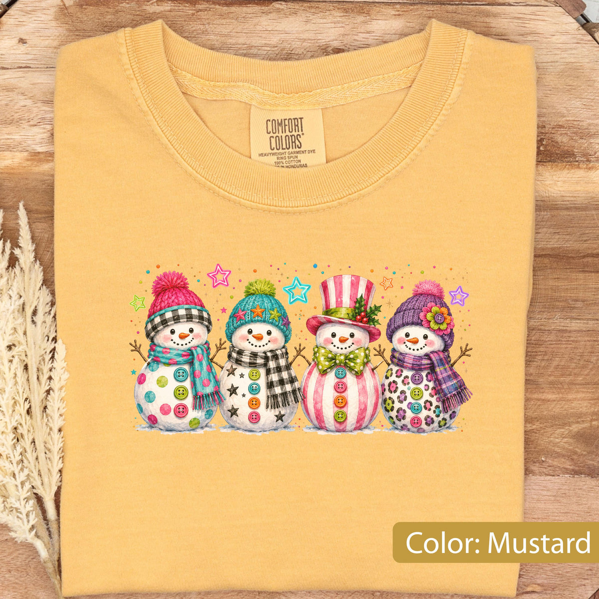 May include: A mustard yellow t-shirt with a graphic print of four snowmen wearing colorful hats and scarves. The snowmen are decorated with buttons and patterns. The shirt features a Comfort Colors label at the neck, indicating it is made of cotton.