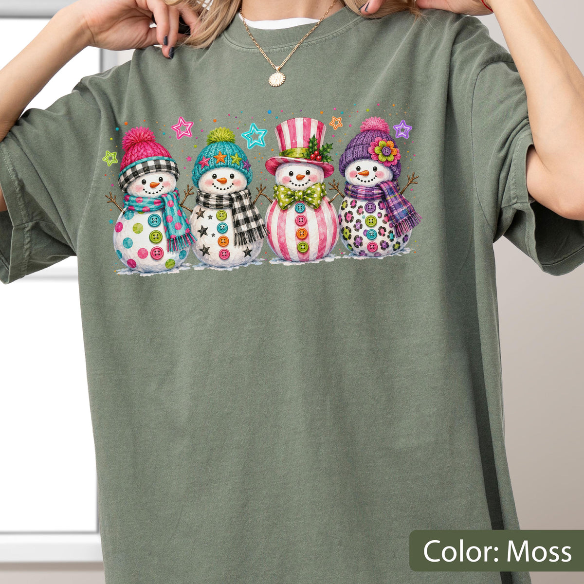 May include: A moss green t-shirt featuring a graphic print of four snowmen standing in a row. Each snowman wears a colorful hat and scarf, with buttons on their bodies. The snowmen are decorated with patterns including plaid, stripes, and leopard print, accented by small colorful stars.