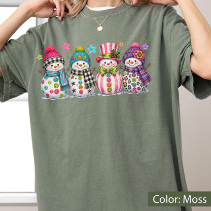 May include: A moss green t-shirt featuring a graphic print of four snowmen standing in a row. Each snowman wears a colorful hat and scarf, with buttons on their bodies. The snowmen are decorated with patterns including plaid, stripes, and leopard print, accented by small colorful stars.