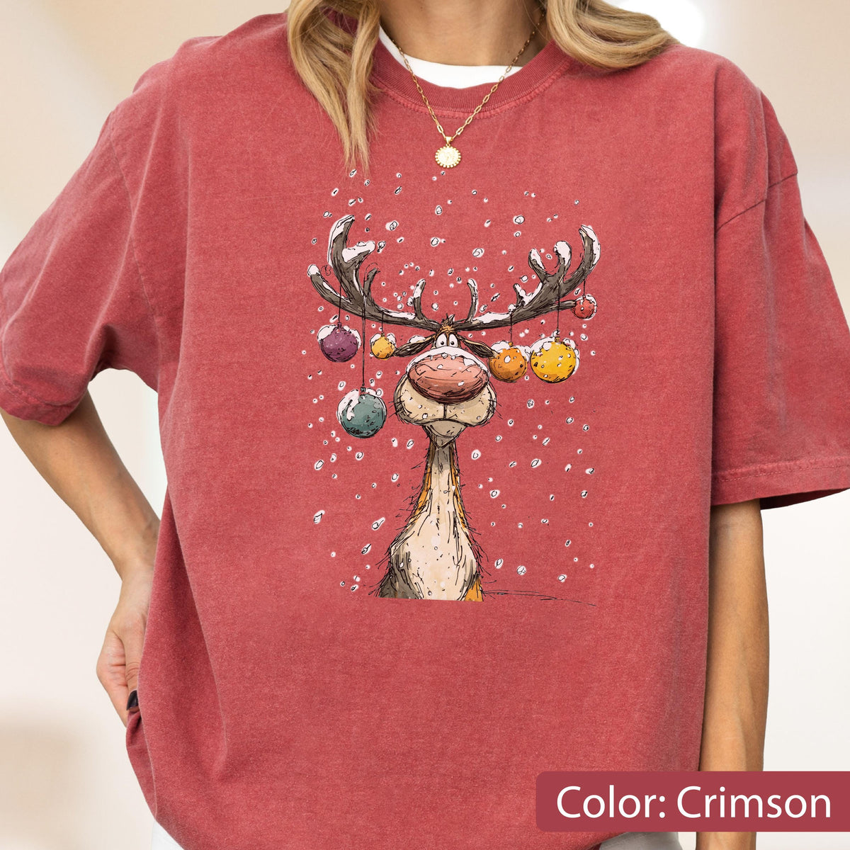 May include: A crimson short-sleeve t-shirt featuring a graphic illustration of a reindeer with large antlers decorated with colorful hanging ornaments. The reindeer is depicted against a background of falling white snow, creating a festive scene.