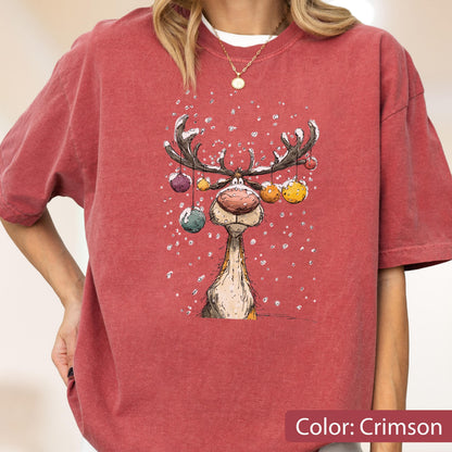May include: A crimson short-sleeve t-shirt featuring a graphic illustration of a reindeer with large antlers decorated with colorful hanging ornaments. The reindeer is depicted against a background of falling white snow, creating a festive scene.