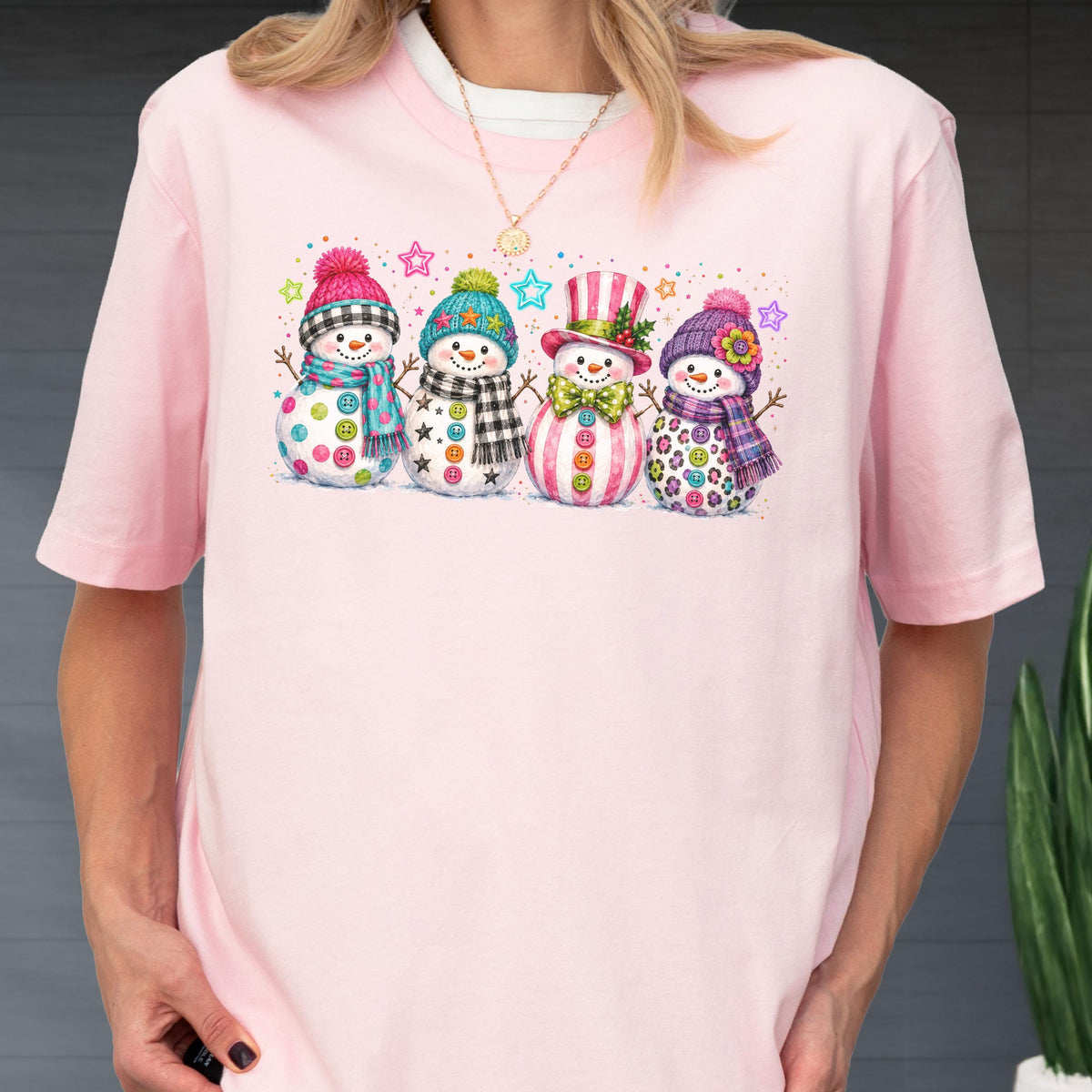 May include: A light pink short-sleeve t-shirt featuring a graphic print of four snowmen standing in a row. Each snowman wears a colorful hat and scarf with unique patterns, including stripes, plaid, and polka dots, accented by small stars.