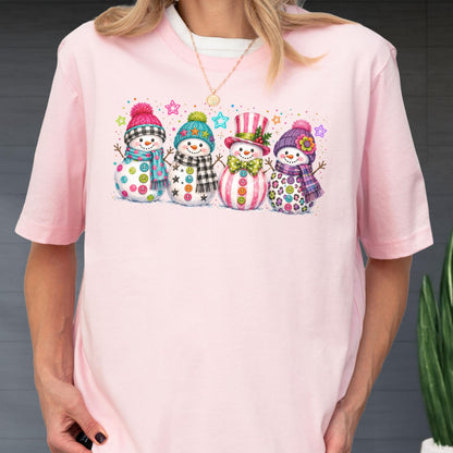 May include: A light pink short-sleeve t-shirt featuring a graphic print of four snowmen standing in a row. Each snowman wears a colorful hat and scarf with unique patterns, including stripes, plaid, and polka dots, accented by small stars.