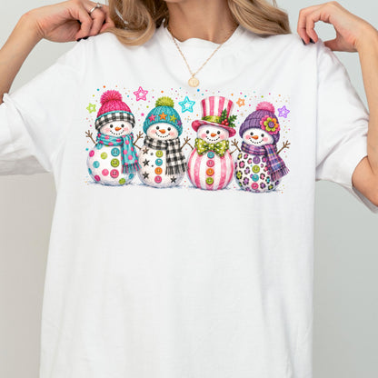 May include: A white t-shirt featuring a graphic of four snowmen standing in a row. Each snowman wears a colorful hat and scarf, with buttons on their bodies. The design includes small stars and dots in various bright colors above the snowmen.