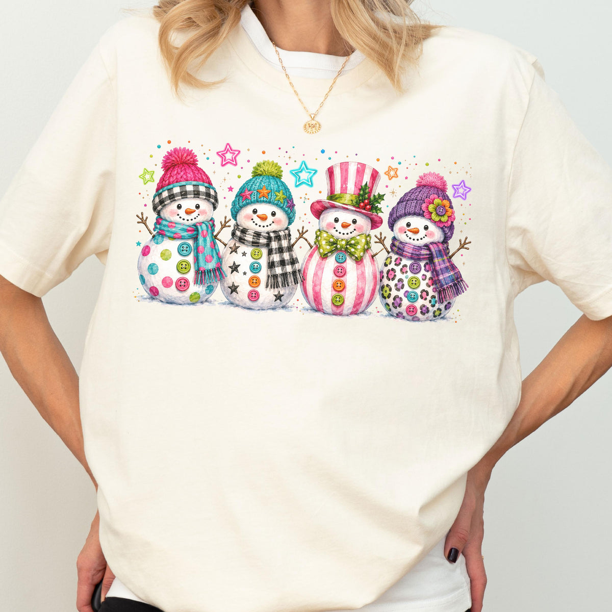 May include: A cream-colored t-shirt featuring a graphic print of four snowmen standing in a row. Each snowman wears a unique hat and scarf, decorated with colorful buttons and surrounded by small, vibrant stars and confetti-like dots.