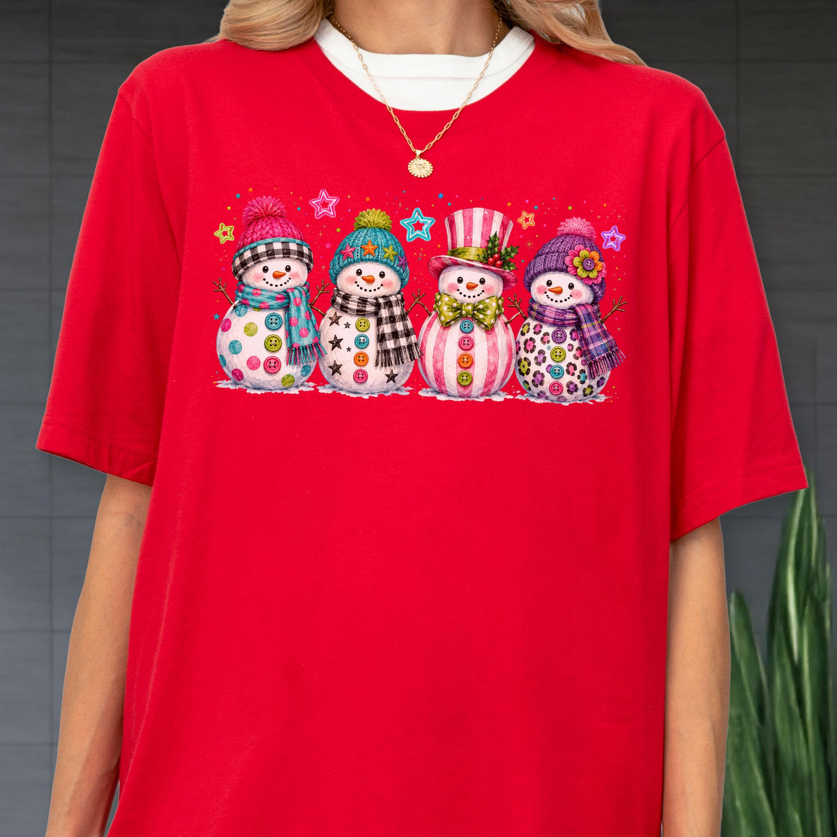 May include: A bright red short-sleeved t-shirt featuring a graphic print of four snowmen standing in a row. Each snowman wears a unique colorful hat and scarf, decorated with various patterns including buttons, stars, and flowers, accented by small stars.