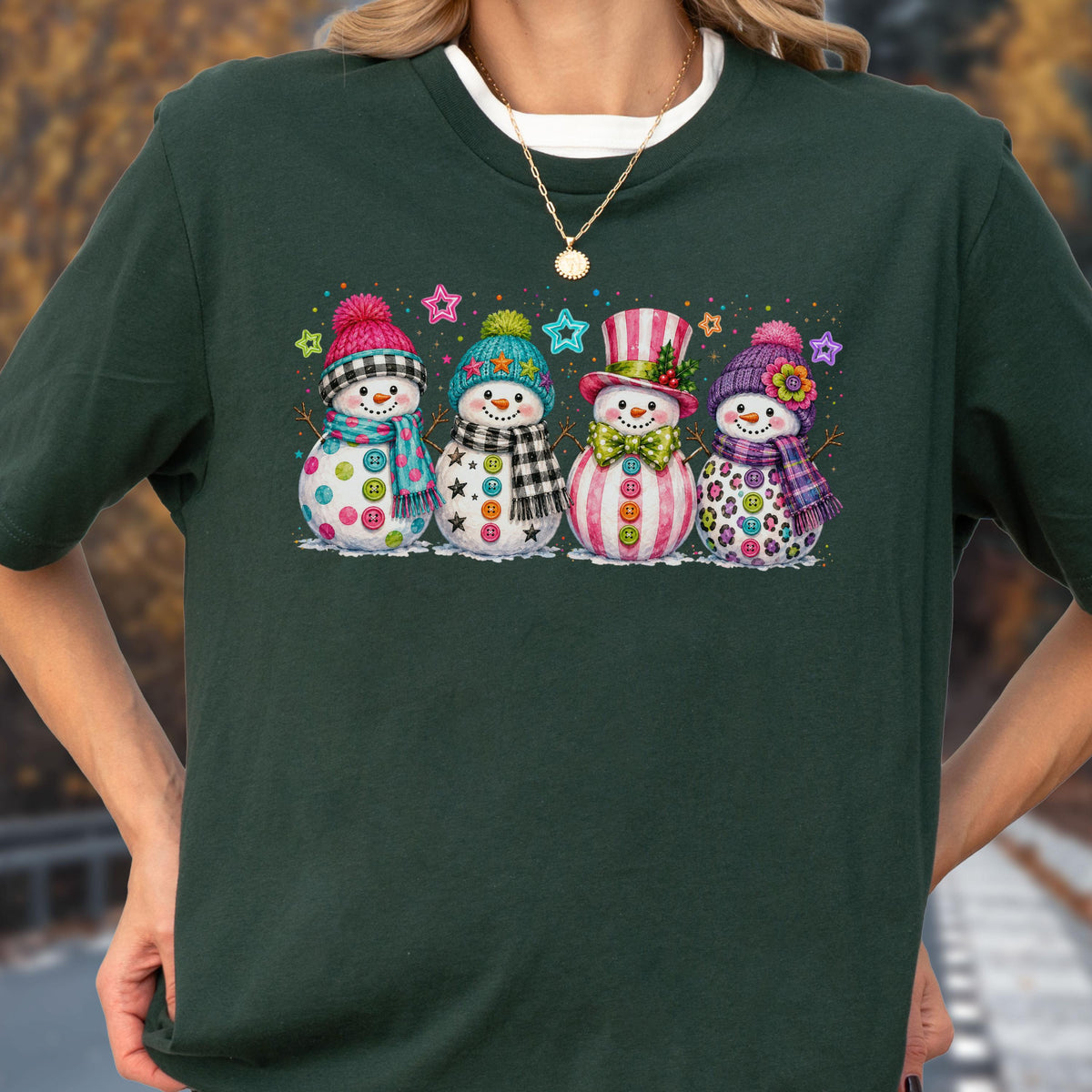 May include: A dark green short-sleeved t-shirt featuring a graphic print of four snowmen standing in a row. Each snowman wears a colorful winter hat and scarf, with buttons on their bodies and small star shapes scattered in the background.