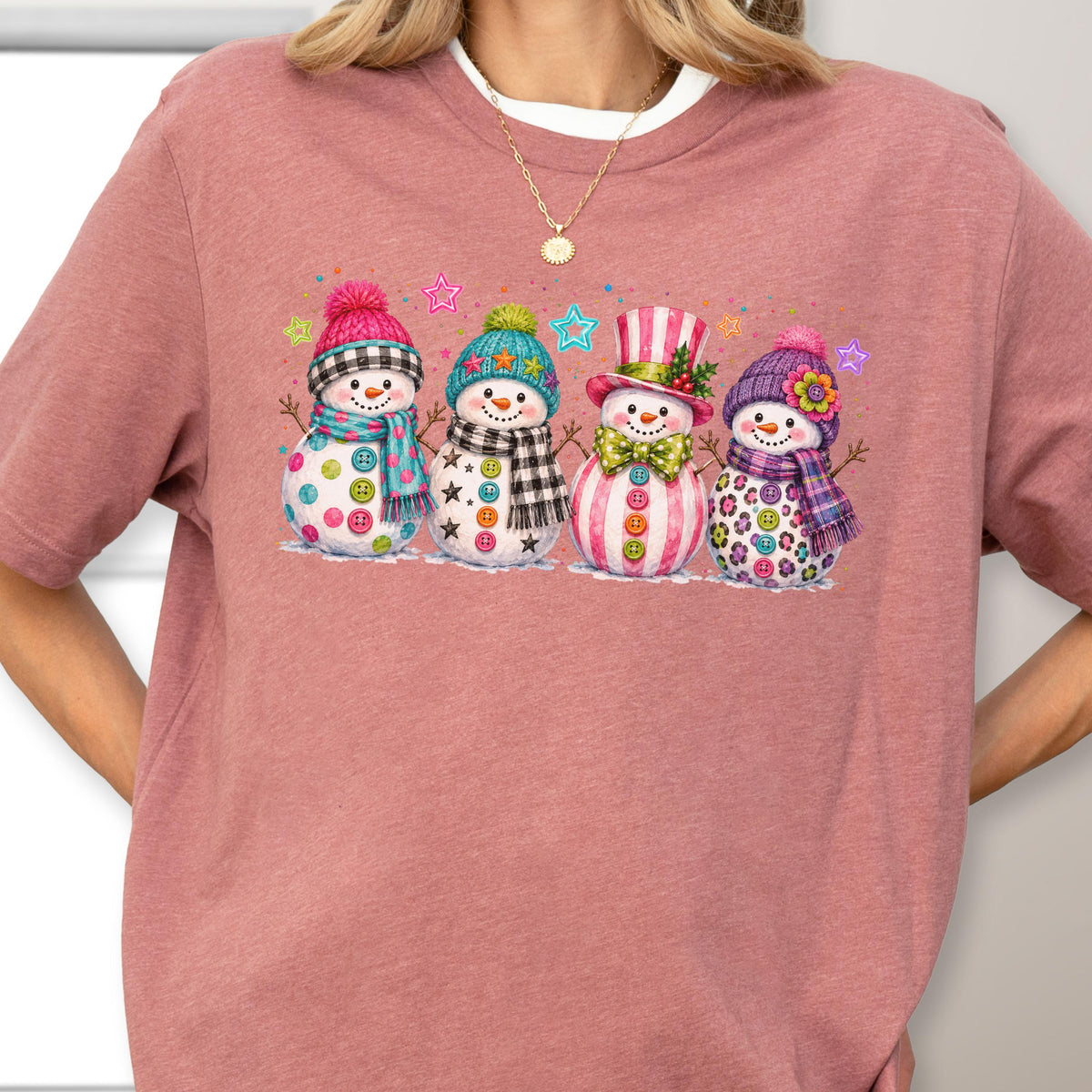 May include: A mauve short-sleeved t-shirt featuring a graphic of four snowmen. The snowmen wear colorful hats, scarves, and button embellishments. The design includes small star accents in blue, pink, and green colors surrounding the snowmen figures.