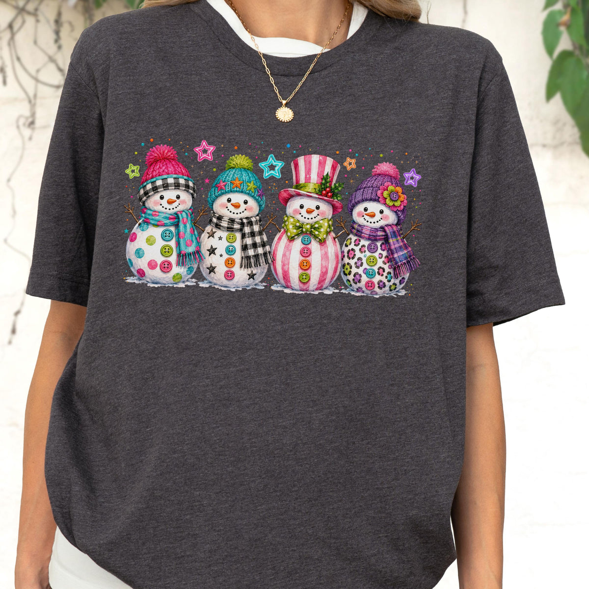 May include: A dark charcoal gray short-sleeve t-shirt featuring a graphic print of four snowmen. Each snowman wears a colorful hat and scarf, with buttons on their bodies and small star shapes scattered above them in various bright colors.