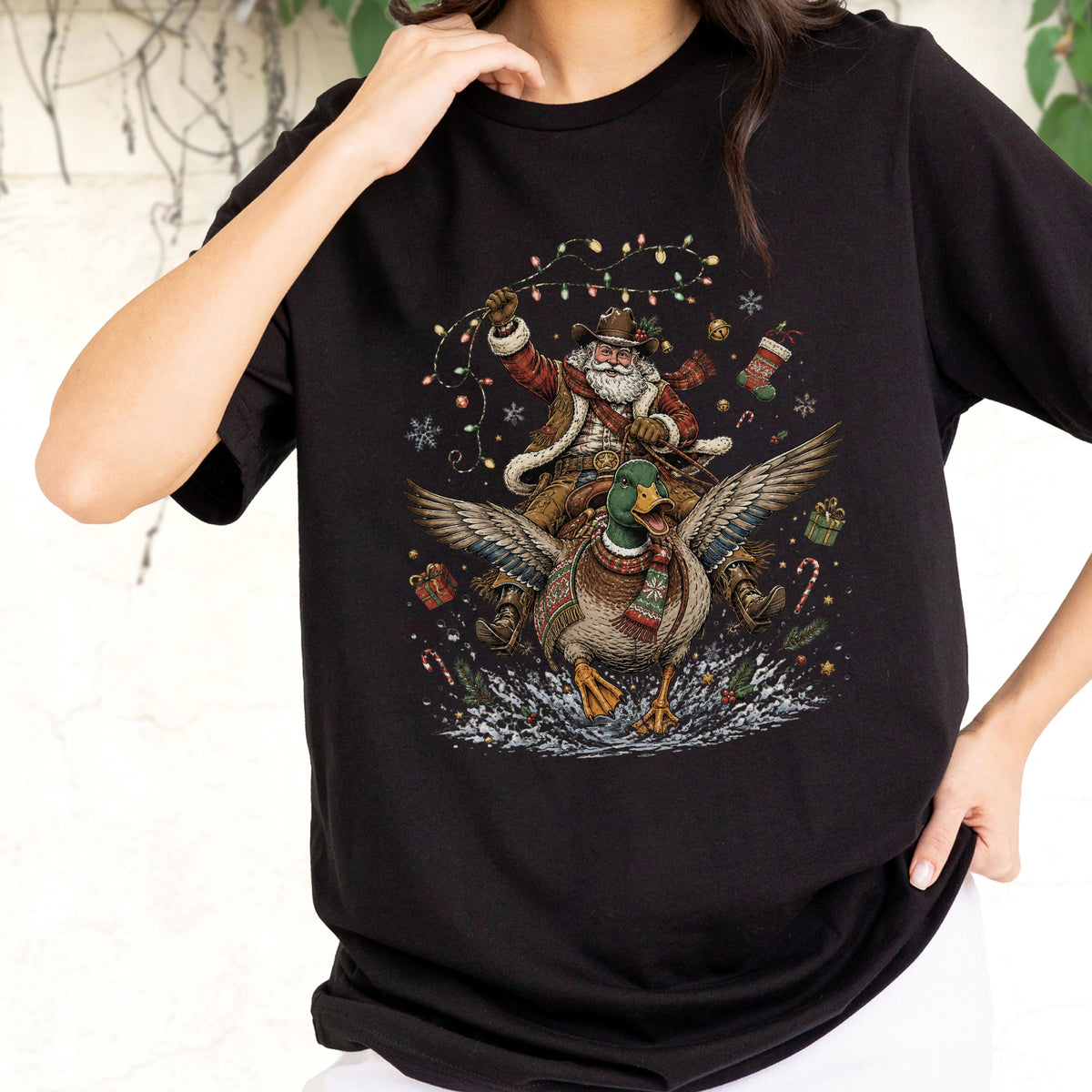 May include: A black t-shirt featuring a graphic print of a cowboy Santa Claus riding a flying mallard duck. The design includes colorful string lights, snowflakes, gift boxes, and candy canes surrounding the central figures in a festive scene.