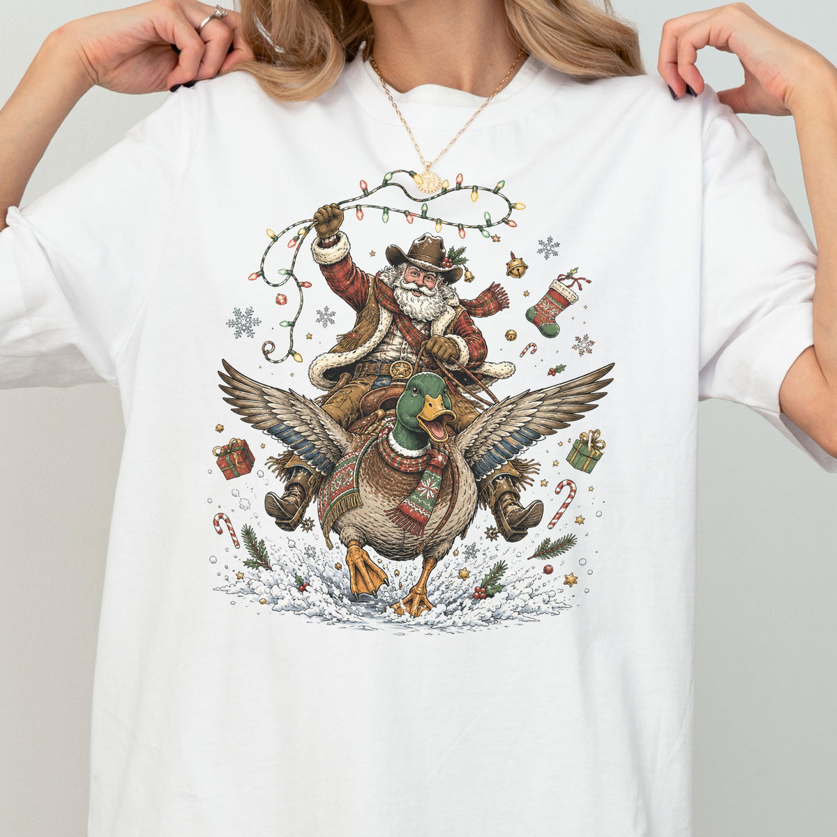 May include: A white t-shirt featuring a graphic illustration of Santa Claus wearing a cowboy hat and vest, riding a mallard duck. The scene includes Christmas lights, a stocking, gift boxes, candy canes, and snowflakes on a snowy ground.